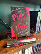 A copy of the book 'Pink Ink' by Avina St. Graves is displayed on a wooden bookshelf, standing upright on a small gold book stand next to other copies of the book.