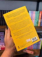 A hand holds the back cover for 'Please Don't Go' by E. Salvador, featuring black text on a yellow background, in front of a blurry bookshelf full of books.