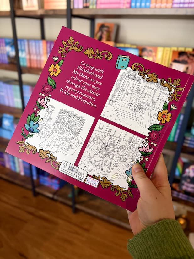 A hand holding the back of a copy of 'Pride and Prejudice Colouring Book' featuring a dark pink background with a floral border and black and white illustrated pages.