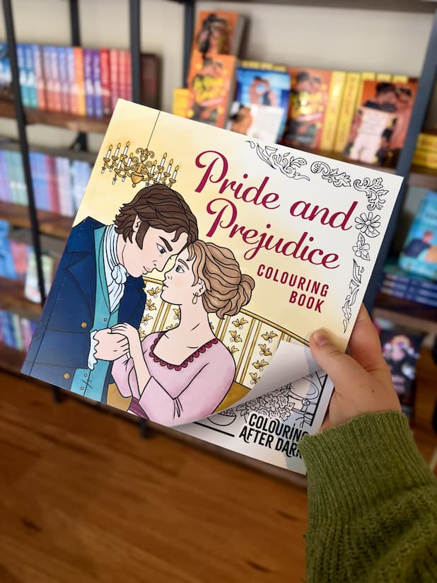 A hand holding a copy of 'Pride and Prejudice Colouring Book' featuring an illustrated Regency couple in an elegant room, with decorative floral borders.