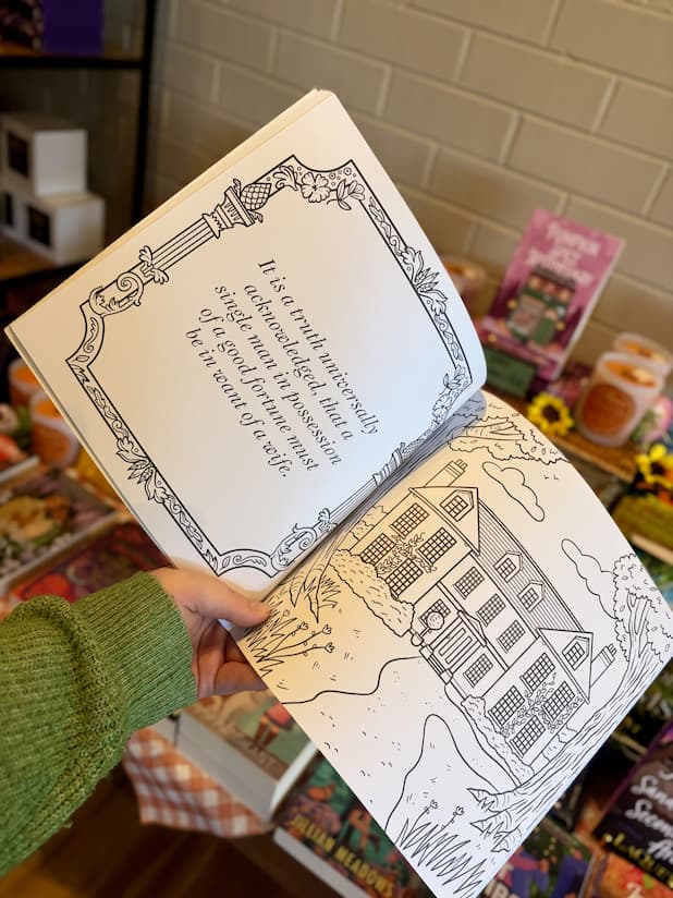 A hand holding an open copy of 'Pride and Prejudice Colouring Book' featuring an illustrated multistory house and a quote.