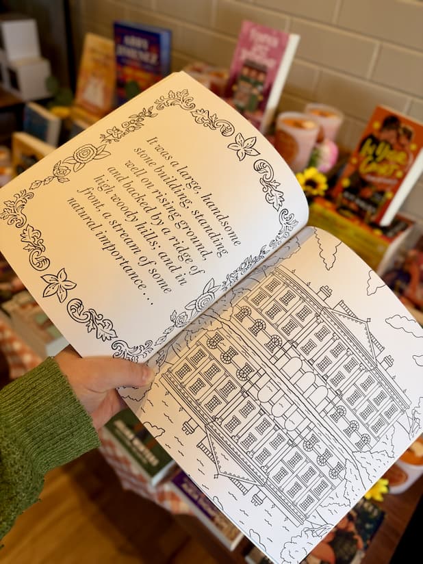 A hand holding an open copy of 'Pride and Prejudice Colouring Book' featuring an illustrated multistory house and a quote.