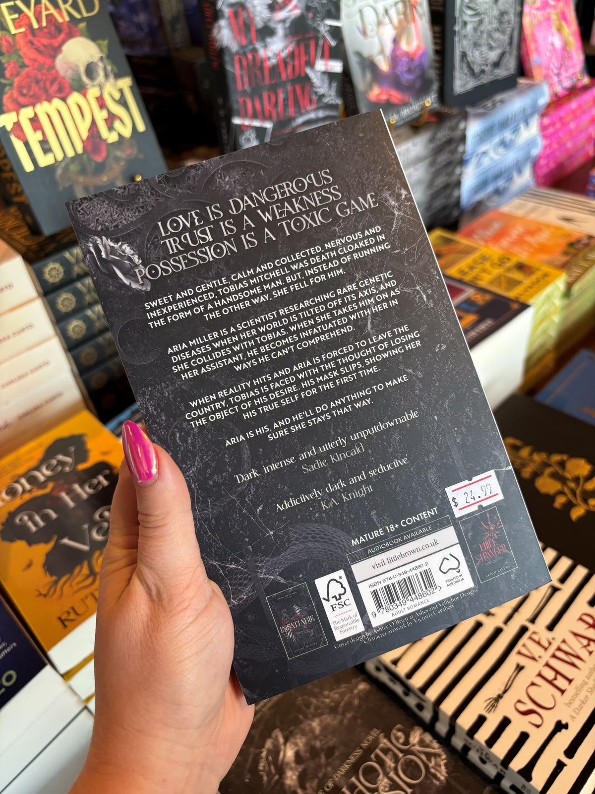 A hand with pink nail polish holds the back cover of 'Psychotic Obsession' by Leigh Rivers in a bookstore with other books blurry in the background. The cover features a dark, marbled background and white text.