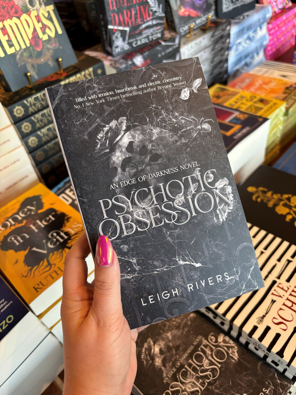 A hand with pink nail polish holds the book cover of 'Psychotic Obsession' by Leigh Rivers in a bookstore with other books blurry in the background. The cover features a dark, marbled background, a decaying skull, and text.