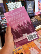 A hand holds the back cover for 'Pumpkin Spice Apothecary' by Melinda Terranova and Cassandra Doon, featuring a whimsical illustration of a pine trees, and a pink autumn-themed background with white font for the blurb.