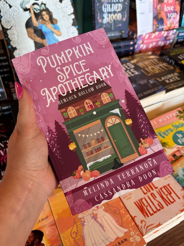 A hand holds the book cover for 'Pumpkin Spice Apothecary' by Melinda Terranova and Cassandra Doon, featuring a whimsical illustration of a green apothecary shop with pumpkins, pine trees, and a pink autumn-themed background. With a table of blurry books in the background. 