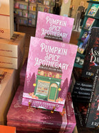 A stack of 'Pumpkin Spice Apothecary' by Melinda Terranova and Cassandra Doon, featuring a whimsical illustration of a green apothecary shop with pumpkins, pine trees, and a pink autumn-themed background on the cover. With a blurry bookshop in the background.