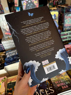 A hand holds the back cover of the special edition version of 'Queen of Faces' by Petra Lord with the blurb in white text on a black background featuring waves, a blue butterfly and a strike of lightning. The book is held in front of a table full of other books blurry in the background.