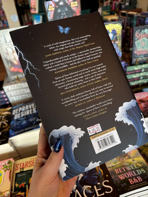 A hand holds the back cover of the special edition version of 'Queen of Faces' by Petra Lord with the blurb in white text on a black background featuring waves, a blue butterfly and a strike of lightning. The book is held in front of a table full of other books blurry in the background.
