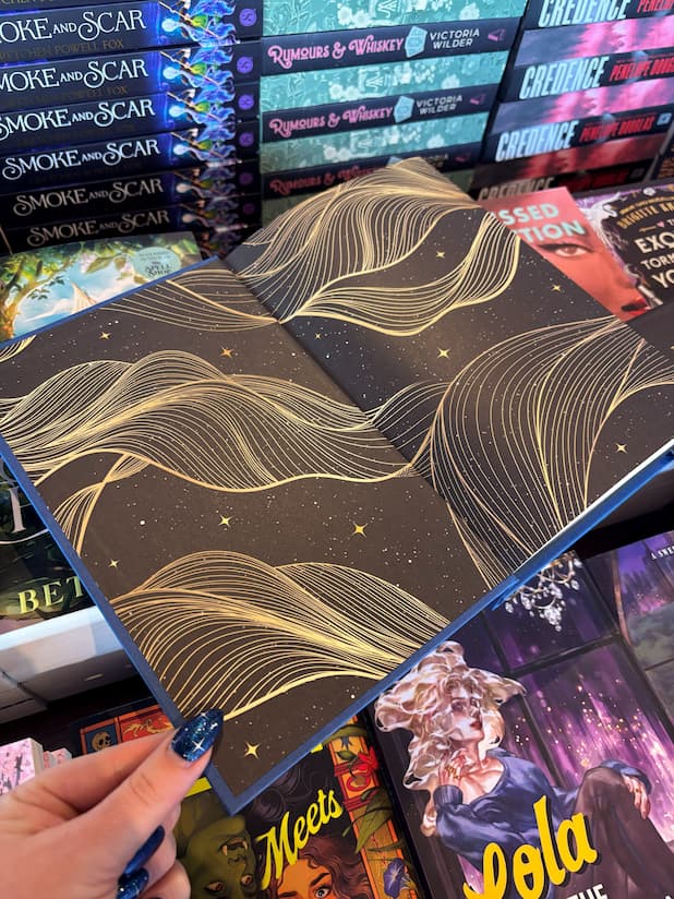 A hand holds the open the book cover of the special edition version of 'Queen of Faces' by Petra Lord showing the end pages that have a black starry design with golden wavy elements. The book is held in front of a table full of other books blurry in the background.