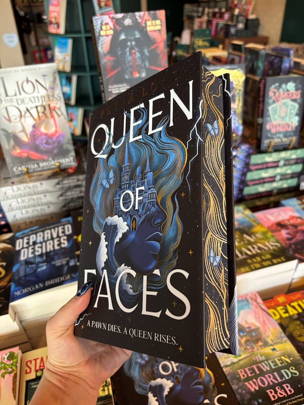 A hand holds the book cover of the special edition version of 'Queen of Faces' by Petra Lord with a face that has the top half morphing into a castle and wavy design on a dark starry background. The edges are black with a holden wavy design and two blue butterflies. The book is held in front of a table full of other books blurry in the background.