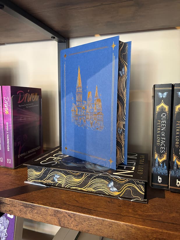 A copy of the special edition version of 'Queen of Faces' by Petra Lord with the dust jacket removed shows a gold foiled castle on a blue hardcover, sitting on top of another copy of the book with digitally sprayed edged facing out. The book sits on a wooden shelf in front of a cream coloured wall. 