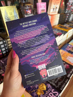 A hand with pink nail polish holds a the back cover of 'Reckless Vow' by Gemma Morr in a bookstore setting, with other books visible in the background. The cover features a dark purple and black design with pink and purple mountain imagery and white text.