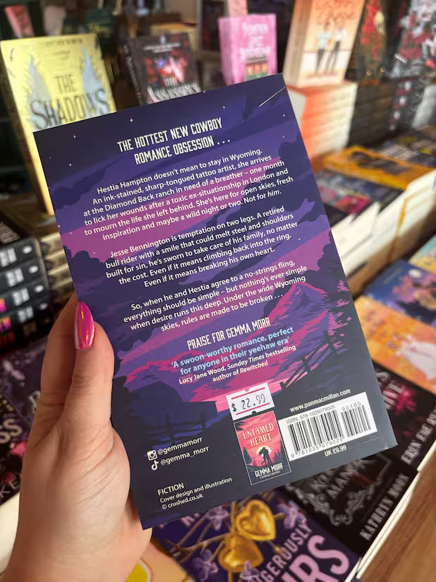 A hand with pink nail polish holds a the back cover of 'Reckless Vow' by Gemma Morr in a bookstore setting, with other books visible in the background. The cover features a dark purple and black design with pink and purple mountain imagery and white text.