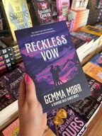 A hand with pink nail polish holds a paperback copy of the book 'Reckless Vow' by Gemma Morr, set against a background of bookshelves filled with other novels.