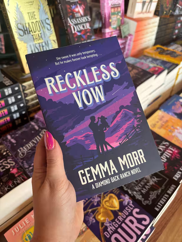 A hand with pink nail polish holds a paperback copy of the book 'Reckless Vow' by Gemma Morr, set against a background of bookshelves filled with other novels.