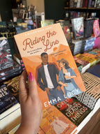 A hand with pink nail polish holding a copy of 'Riding the Line' by Emma Lucy, featuring a man in a leather jacket and a woman in a cowboy hat against an orange landscape.
