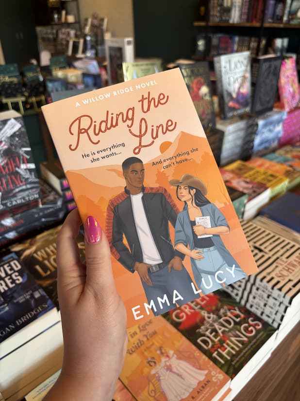 A hand with pink nail polish holding a copy of 'Riding the Line' by Emma Lucy, featuring a man in a leather jacket and a woman in a cowboy hat against an orange landscape.