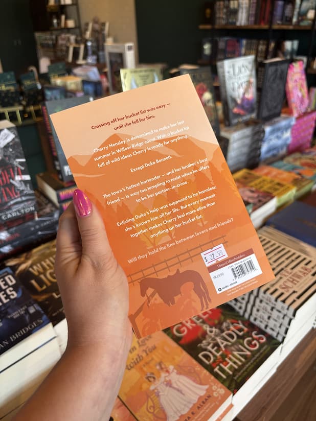 A hand with pink nail polish holding the back of a copy of 'Riding the Line' by Emma Lucy, featuring an orange background, a horse and a motorcycle silhouette, and white text.