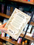 A hand holding the back cover of 'Riftborne' by Grenwich & Lennox. The book cover features a white background with a green snake wrapped around a sword and ornate gold detailing.