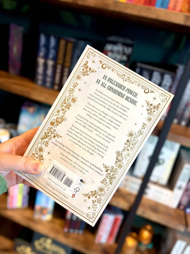 A hand holding the back cover of 'Riftborne' by Grenwich & Lennox. The book cover features a white background with a green snake wrapped around a sword and ornate gold detailing.