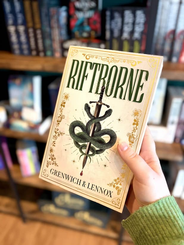 A hand holding a paperback book titled 'Riftborne' by Grenwich & Lennox, featuring a cream cover with a green snake wrapped around a dagger, set against a blurred bookshelf background.