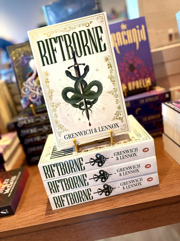 A stack of three 'Riftborne' books by Grenwich & Lennox displayed on a wooden shelf, with the top book propped upright on a small gold stand. The book cover features a white background with a green snake wrapped around a sword and ornate gold detailing.