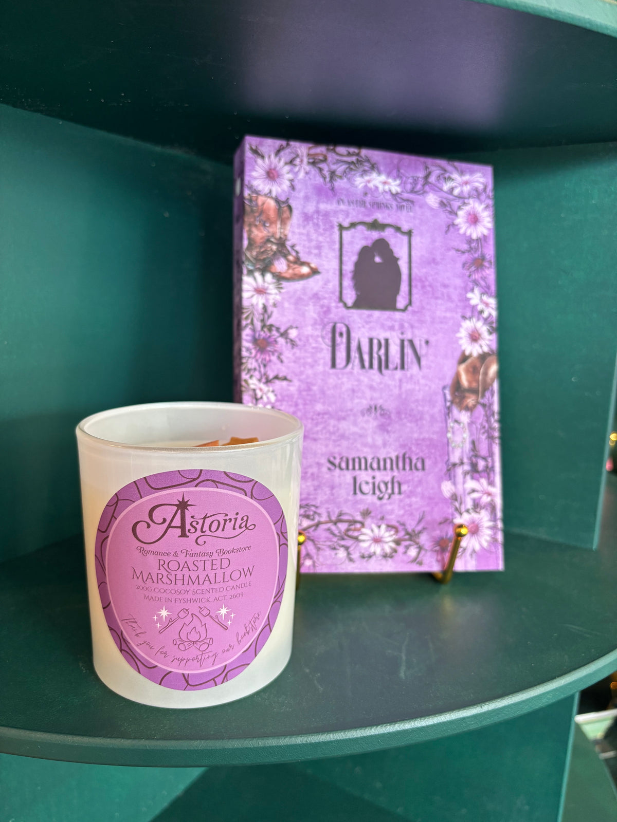 A white candle jar with a purple circular label reading 'Roasted Marshmallow' sits on a dark green shelf next to its inspiration 'Darlin' by Samantha Leigh.