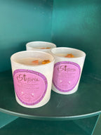Three white soy candles in white glass jars with pink and purple Astoria Roasted Marshmallow labels, displayed on a dark shelf against a teal background.