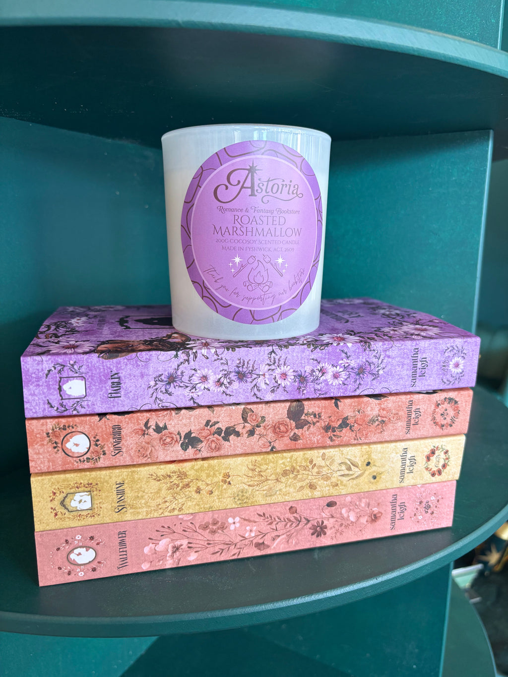 A white scented candle with a lavender circular label reading 'Astoria Roasted Marshmallow' sits on top of a stack of the Aster Springs series by Samantha Leigh which inspired the candle, displayed on a teal shelf.