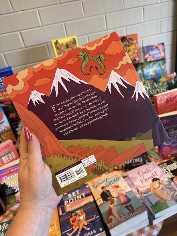 A hand with pink nail polish holds a copy of 'Romantasy Coloring Book' with an illustration of a mountain landscape and white text.