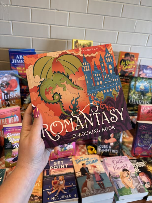 A hand with pink nail polish holds a copy of the 'Romantasy Coloring Book' with a dragon and castle design.