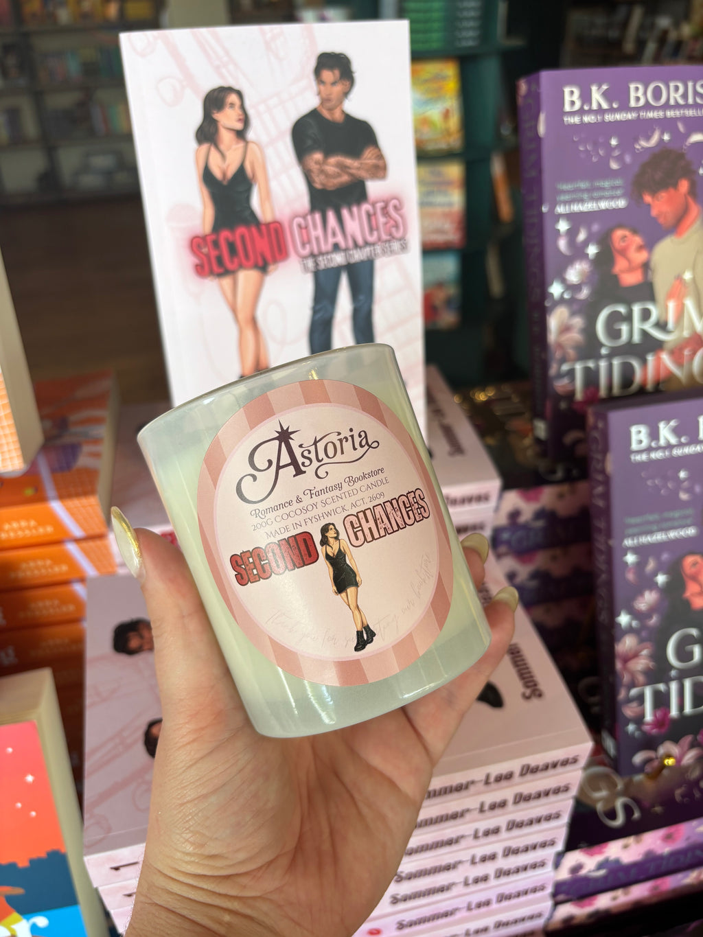 A hand holds a frosted glass candle in front of a display of books, including a prominent 'Second Chances' book by Astoria. The candle features a matching pink and white label with illustrations.