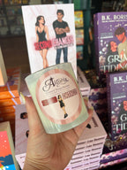 A hand holds a frosted glass candle in front of a display of books, including a prominent 'Second Chances' book by Astoria. The candle features a matching pink and white label with illustrations.