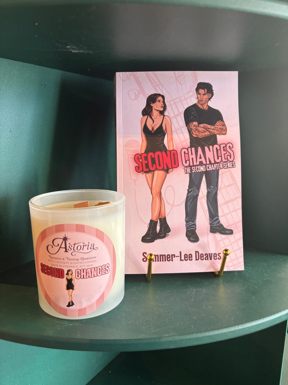 A white jar candle and a pink book titled 'Second Chances' by Summer-Lee Deaves are displayed on a dark teal shelf.