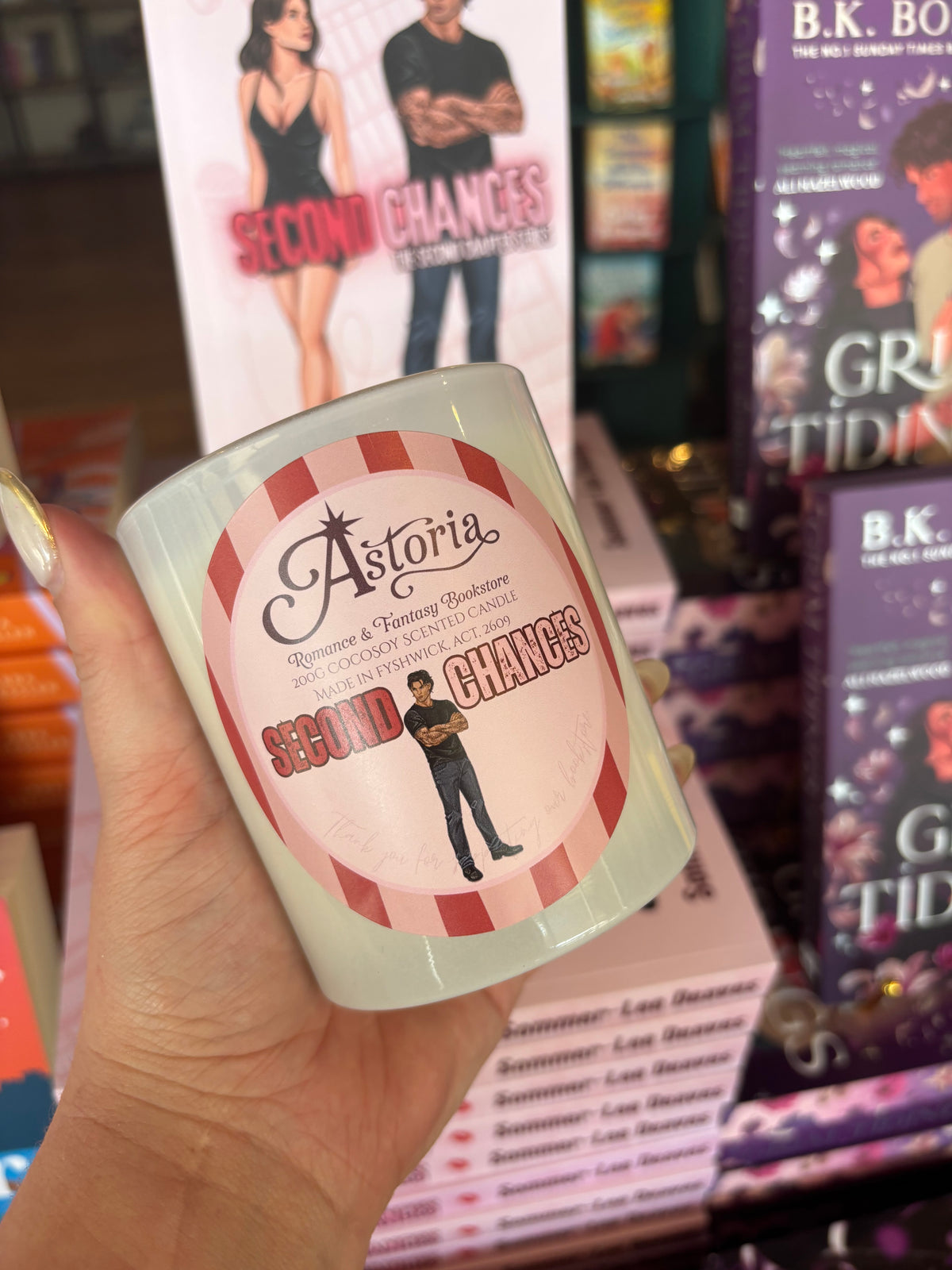 A hand holding a white ceramic candle jar with a pink and red striped label that reads 'Astorla Second Chances.' The background shows a retail or bookstore-like environment with other books and displays visible.