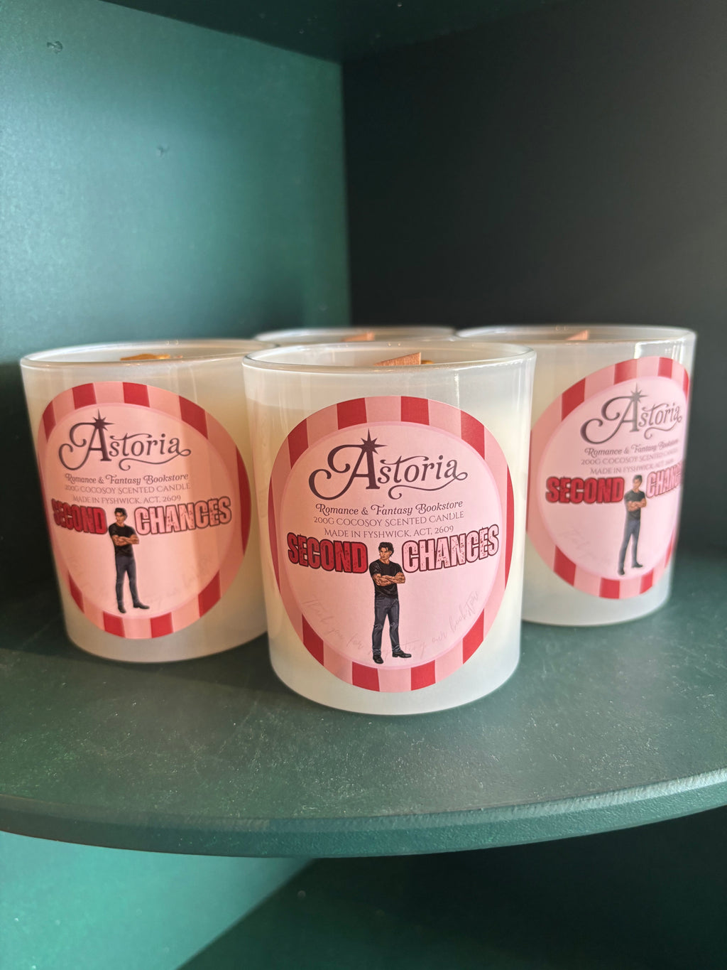 Three white glass jar candles with pink and red labels reading 'Astoria Second Chances' sit on a dark teal shelf against a dark background.