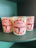 Three white glass jar candles with pink and red labels reading 'Astoria Second Chances' sit on a dark teal shelf against a dark background.