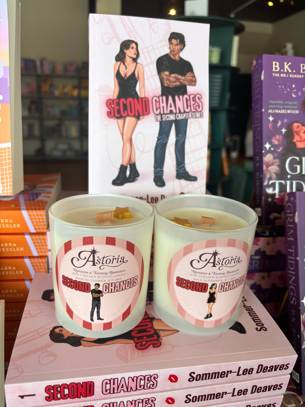Two 'Astoria Second Chances' soy candles by Sommer-Lee Deaves displayed on top of their book packaging, with a matching book cover standing behind them in a retail setting.
