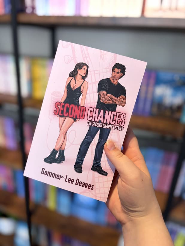 A hand holding a copy of 'Second Chances' by Sommer-Lee Deaves, featuring an illustration of a man and a woman looking over their shoulder at each other with a pale pink background.