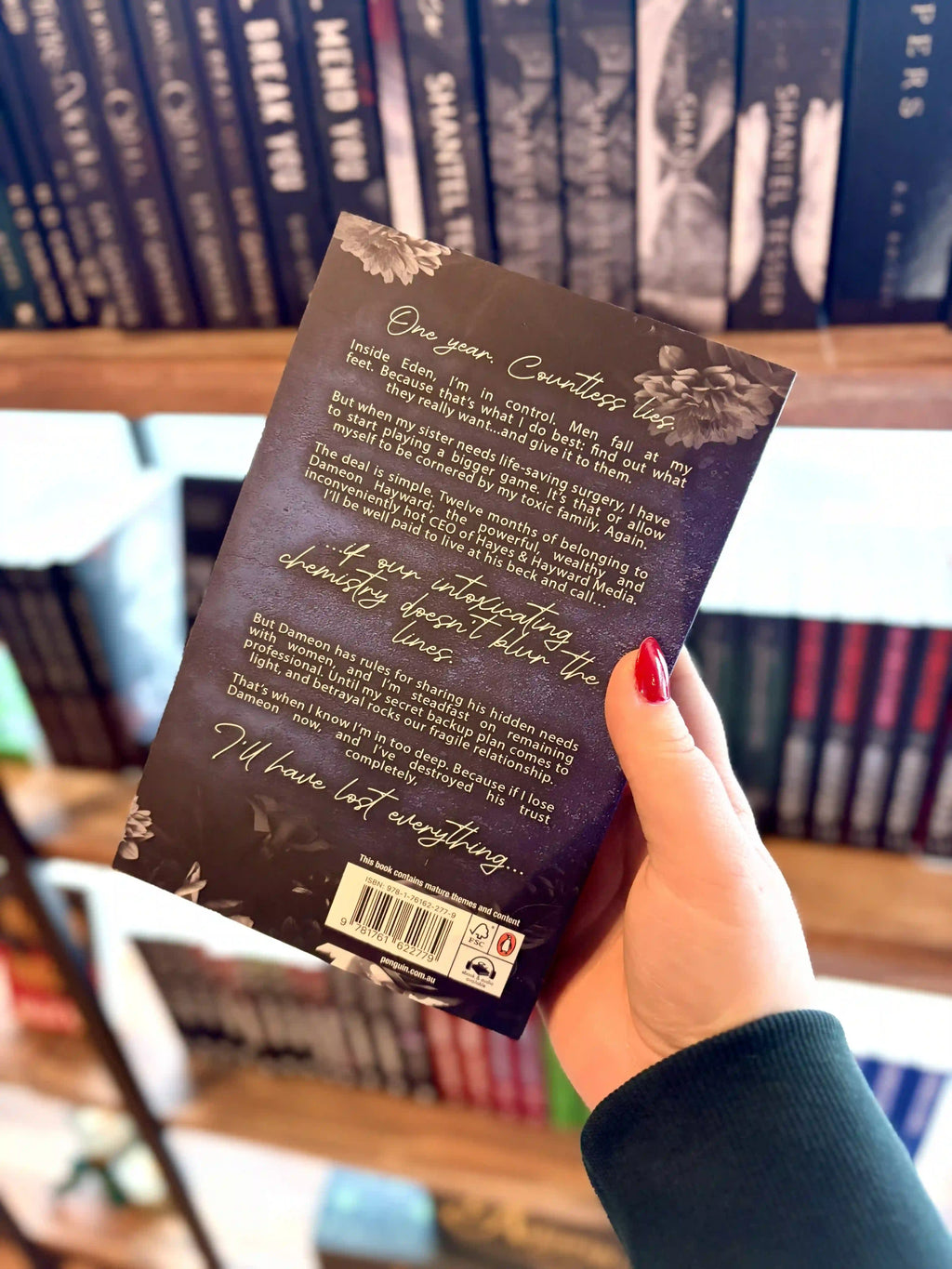 A hand with red nail polish holds the paperback copy of 'Seduced by Eden' by Jade May with the back cover to the camera. Book cover of 'Seduced by Eden' by Jade May. The cover is dark blue with a silver chess piece in the centre and grey roses on the top and bottom of the cover.