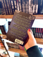 A hand with red nail polish holds the paperback copy of 'Seduced by Eden' by Jade May with the back cover to the camera. Book cover of 'Seduced by Eden' by Jade May. The cover is dark blue with a silver chess piece in the centre and grey roses on the top and bottom of the cover.
