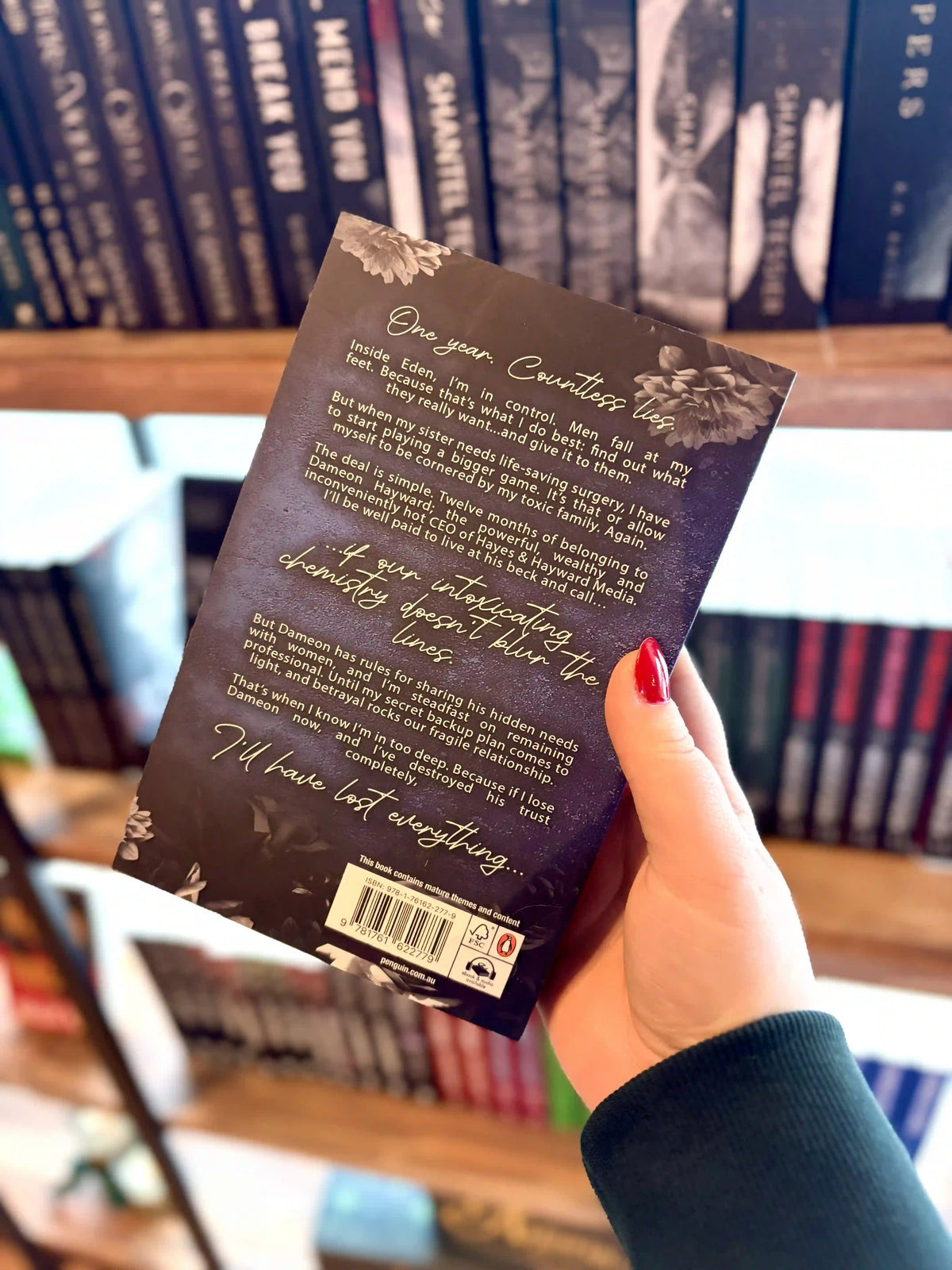 A hand with red nail polish holds the paperback copy of 'Seduced by Eden' by Jade May with the back cover to the camera. Book cover of 'Seduced by Eden' by Jade May. The cover is dark blue with a silver chess piece in the centre and grey roses on the top and bottom of the cover.