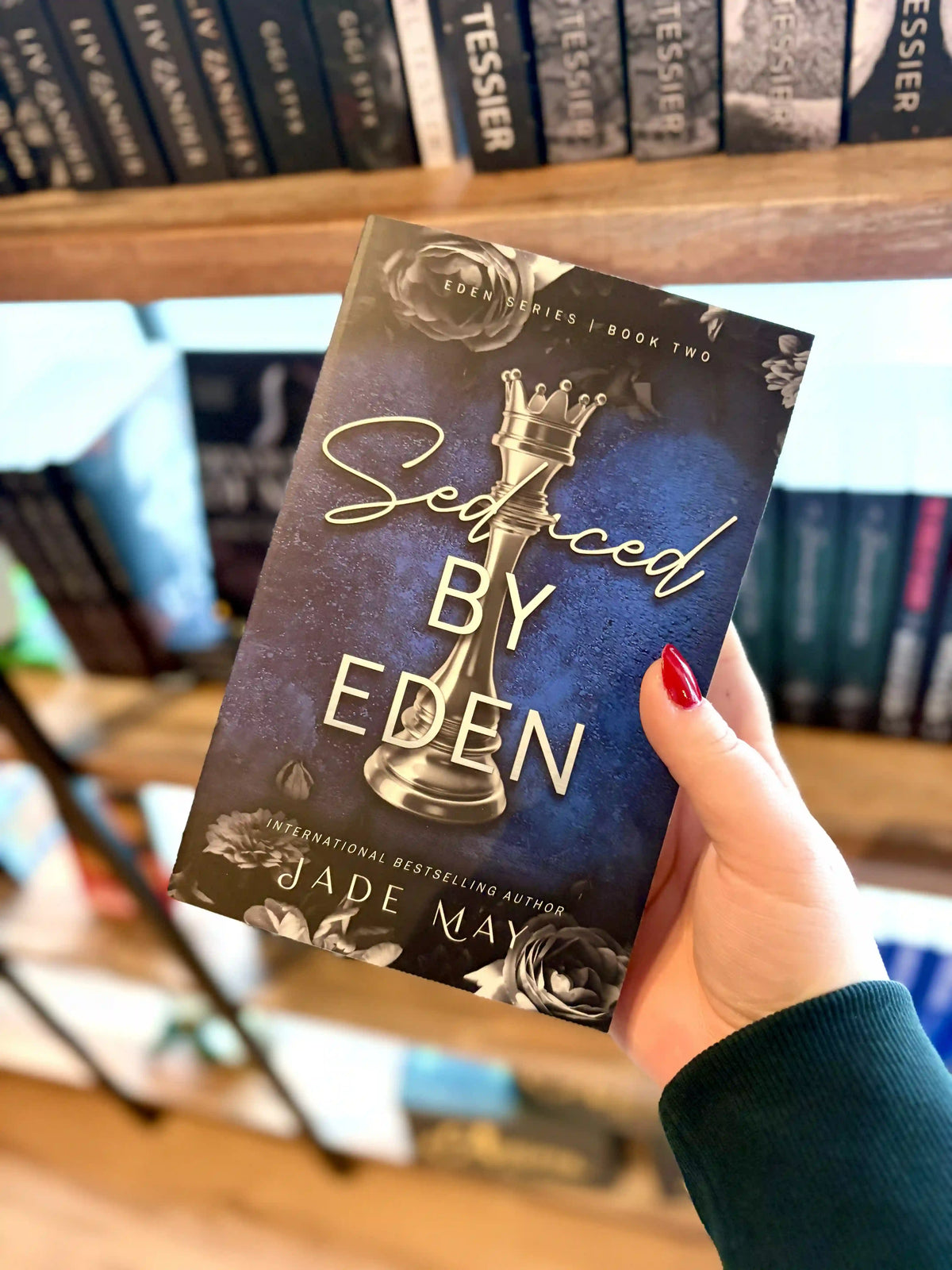A hand with red nail polish holds a paperback copy of the book 'Settled by Eden' by Jade May. The cover is dark blue with a silver chess piece in the centre and grey roses on the top and bottom of the cover.