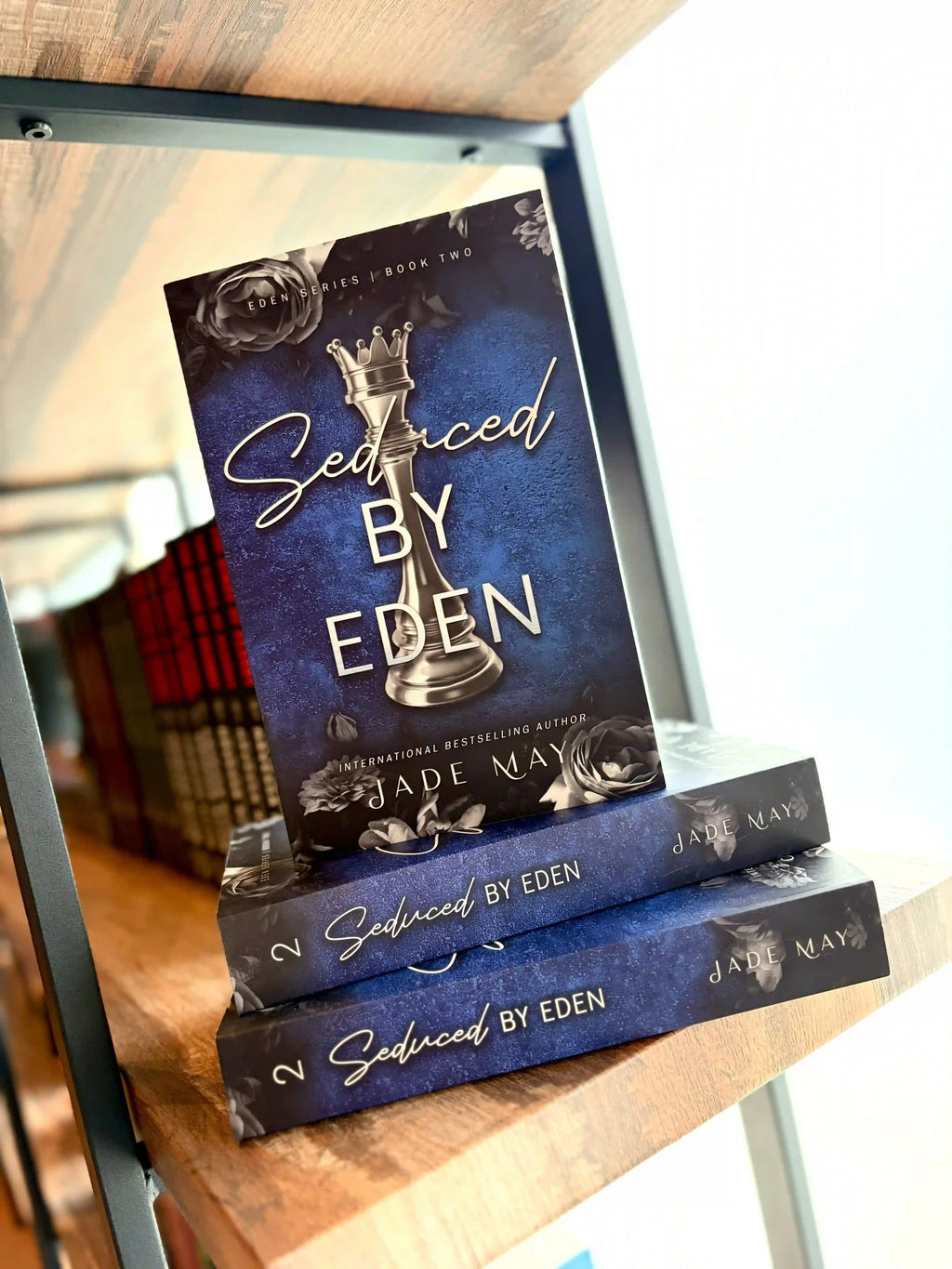 Three copies of the book 'Seduced by Eden' by Jade Maye are stacked on a wooden bookshelf. Book cover of 'Seduced by Eden' by Jade May. The cover is dark blue with a silver chess piece in the centre and grey roses on the top and bottom of the cover.