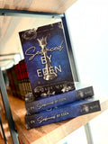 Three copies of the book 'Seduced by Eden' by Jade Maye are stacked on a wooden bookshelf. Book cover of 'Seduced by Eden' by Jade May. The cover is dark blue with a silver chess piece in the centre and grey roses on the top and bottom of the cover.