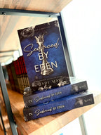 Three copies of the book 'Seduced by Eden' by Jade Maye are stacked on a wooden bookshelf. Book cover of 'Seduced by Eden' by Jade May. The cover is dark blue with a silver chess piece in the centre and grey roses on the top and bottom of the cover.