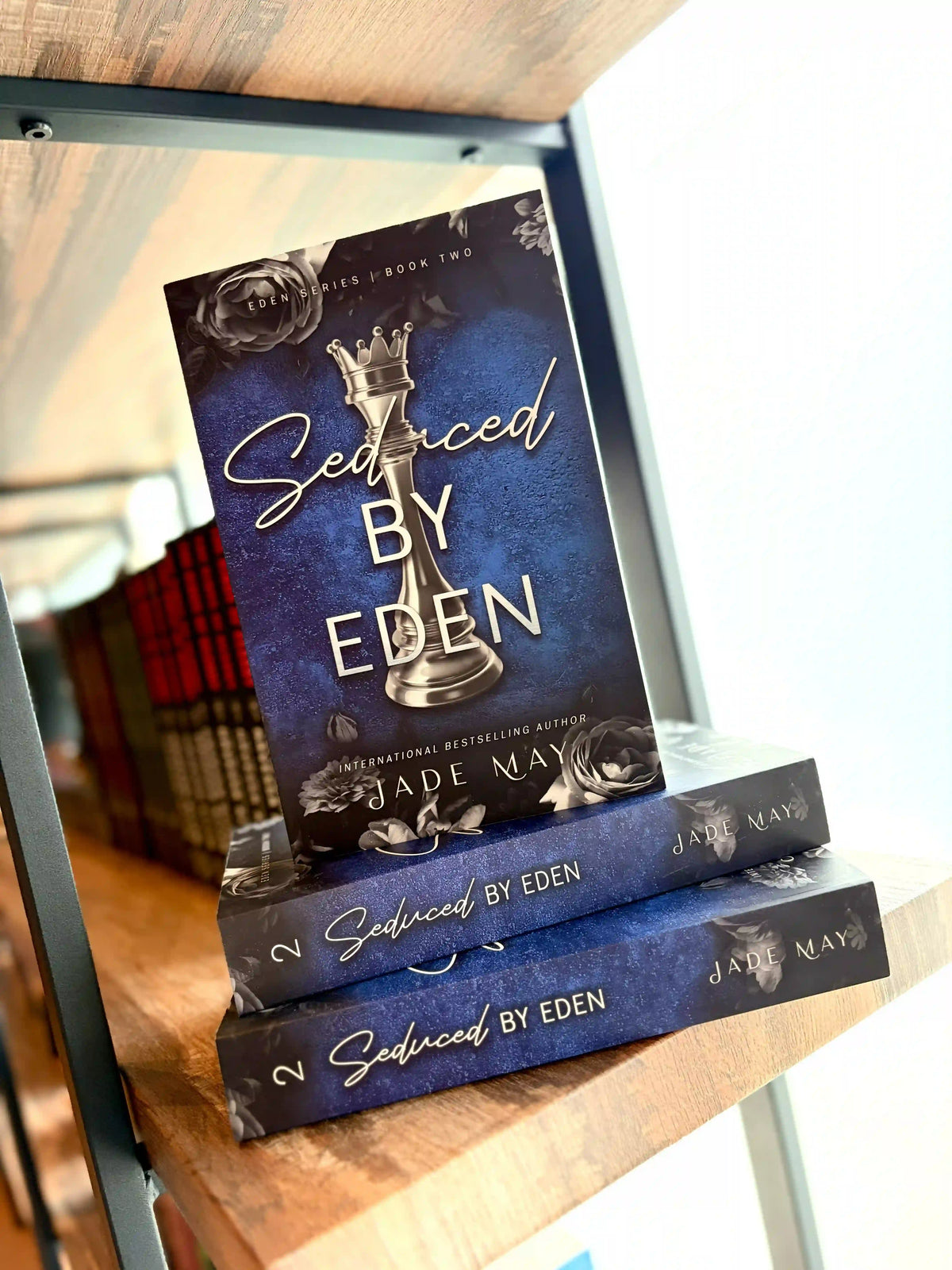 Three copies of the book 'Seduced by Eden' by Jade Maye are stacked on a wooden bookshelf. Book cover of 'Seduced by Eden' by Jade May. The cover is dark blue with a silver chess piece in the centre and grey roses on the top and bottom of the cover.