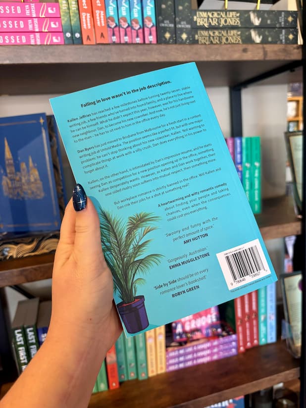 A hand with blue glitter nail polis holds the back cover of the book 'Side by Side' by Jordan Clayden-Lewis in front of a bookshelf. 