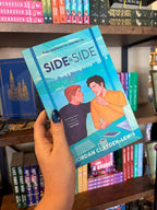 A hand with blue glitter nail polis holds the book 'Side by Side' by Jordan Clayden-Lewis in front of a bookshelf. The cover features an illustrated scene of two men sitting at a desk with a laptop and coffee mug, with a city skyline in the background.